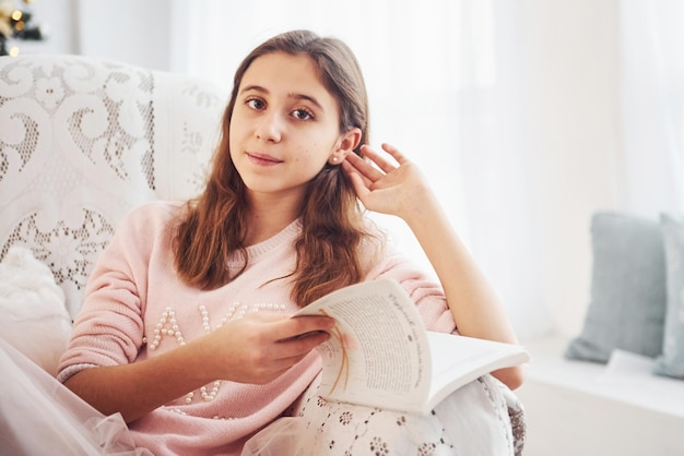 Cute female teen sits indoors in the living room with book at holidays time.