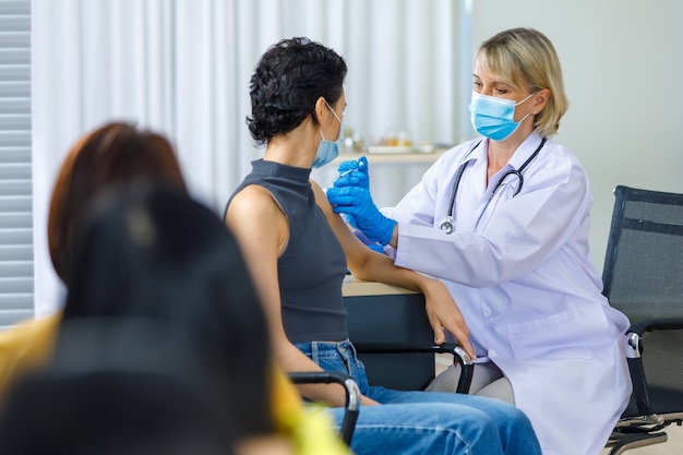 Симпатичная женщина в маске сидит и смотрит в камеру, в то время как кавказская женщина-врач в белом лабораторном халате, синие перчатки и стетоскоп вводит вакцину на плечо, когда другие пациенты ждут в очереди.