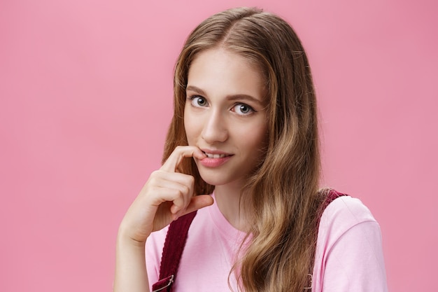 Cute feminine girl learning how make glances and seduce boyfriend with flirty gaze biting finger sensually posing against pink background, flirting making senducing look wearing make-up on date.