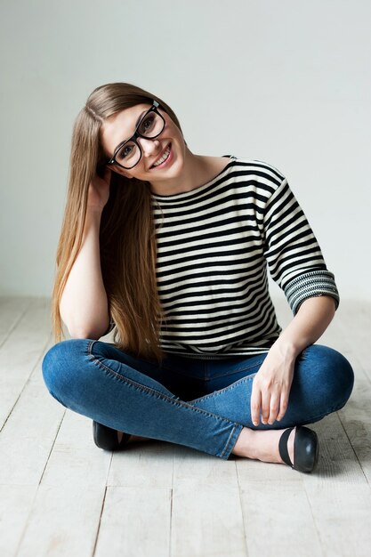 Симпатичный и кокетливый. Красивая молодая женщина в полосатой одежде сидит на деревянном полу и держится за подбородок