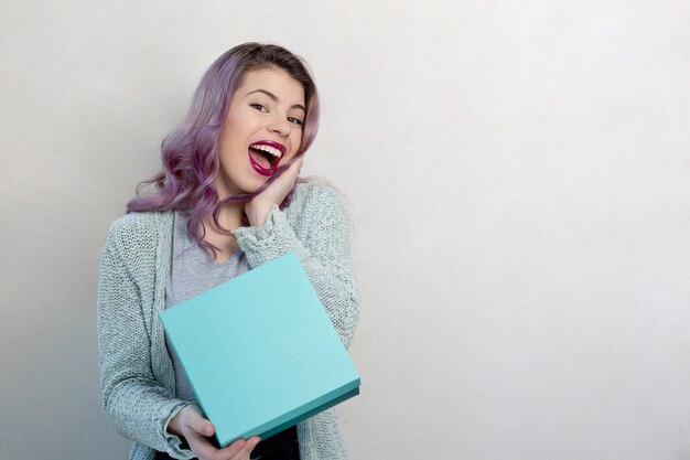 Cute flirty young woman with purple hair and bright makeup holding gift box, studio shot over a grey background. Space for text