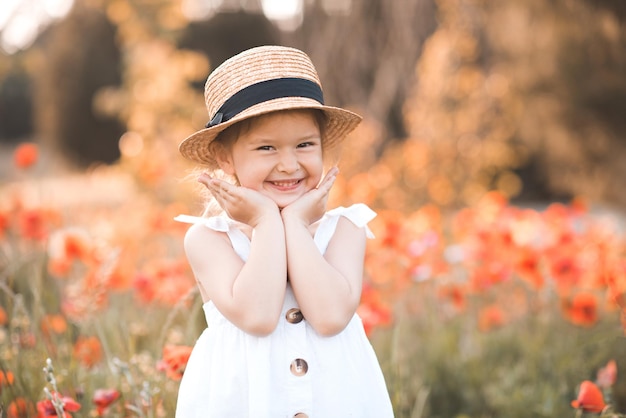 Cute funny baby girl 2-3 year old wear straw hat and white summer dress over flower meadow background. Kid laughing outdoors over nature. Looking at camera. Childhood.