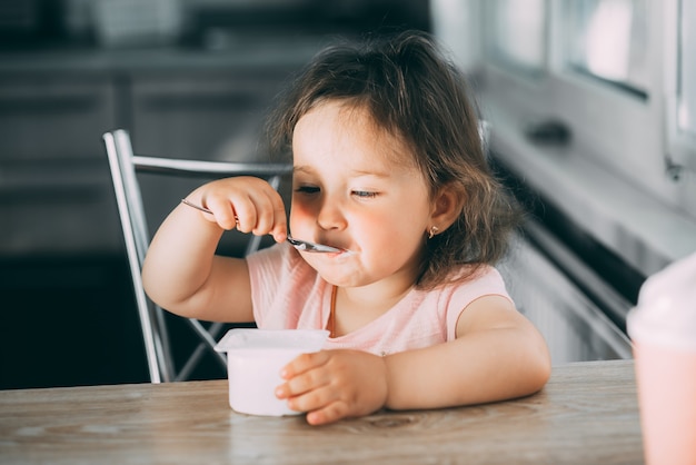 Cute, funny baby girl eating yogurt in the kitchen in a pink dress in the afternoon cute