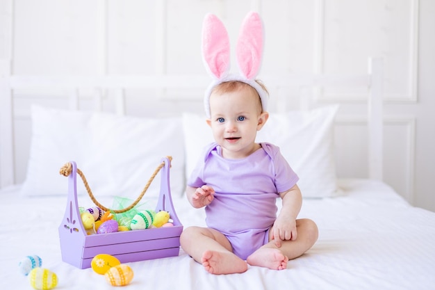 Cute funny baby with bunny ears with colorful Easter eggs and a basket of eggs at home on a bed of white bedding Easter greeting card