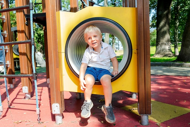 Cute funny blond little young toddler kid child boy crawling climbing through yellow plastic tunnelpipe in playgroundChildren physical development and childhood daycare kindergarten concept