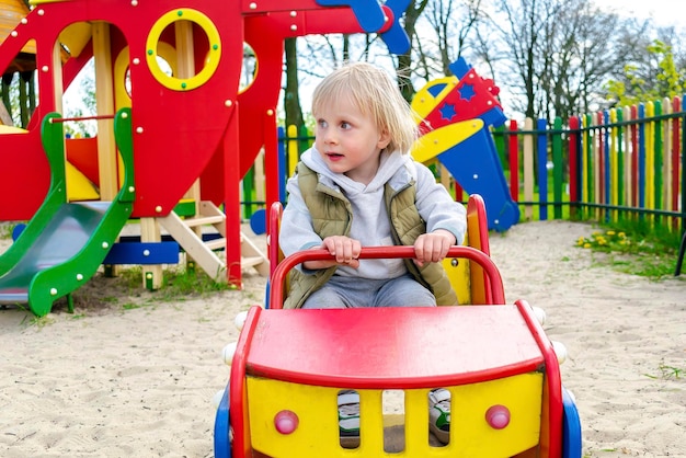 Милый забавный блондин, маленький малыш, ребенок, сидящий и катающийся в игрушечной машине, автомобиль на детской площадке, физическое, эмоциональное развитие детей и концепция детского сада в детском саду