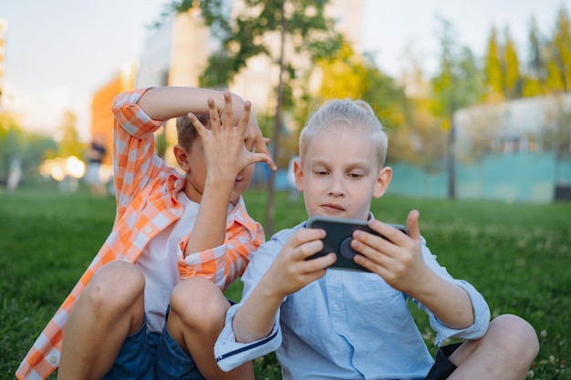 cute funny children shooting selfie with smartphone making frame of fingers. Kids in park sitting on grass having fun together. Image with selective focus