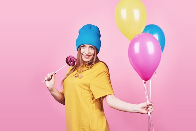 Cute funny girl in blue cap portrait holds an air colorful balloons and lollipop smiling on pink 