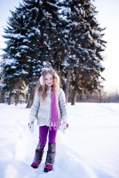 Ragazza carina e divertente del bambino 5-6 anni a piedi nel parco innevato sullo sfondo della natura all'aperto. il bambino indossa giacca invernale, sciarpa e cuffie all'esterno. infanzia.