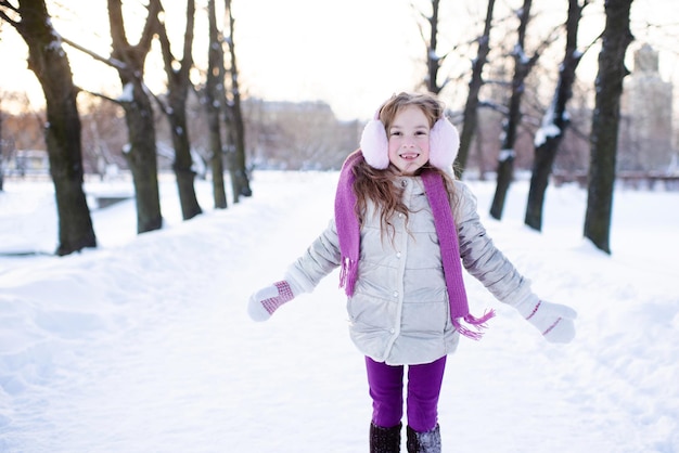 5〜6歳のかわいい面白い子供の女の子は、屋外の自然の背景の上に雪の公園を散歩します。小さな子供は外で冬のジャケット、スカーフ、ヘッドフォンを着用します。子供時代。