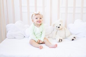 Cute funny little baby girl shows tongue in a crib on a white cotton bed in a green bodysuit in a children's bright room and smiles