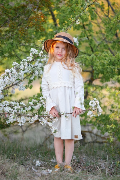 cute funny little blonde girl in a straw hat near a flowering tree in spring laughing