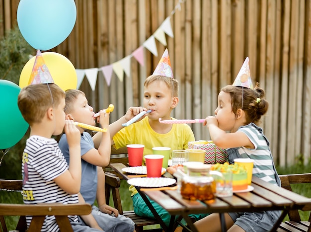 Cute funny nine year old boy celebrating his birthday with\
family or friends with homemade baked cake in a backyard birthday\
party kids wearing party hats and blowing whistles