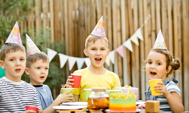 Photo cute funny nine year old boy celebrating his birthday with family or friends with homemade baked cake in a backyard birthday party kids wearing party hats holding paper caps with juice