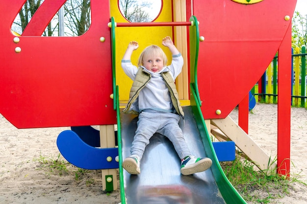 Cute funny smiling blonde little young toddler kid child boy going down slide in playground Children physicalemotional development and childhood daycare kindergarten concept