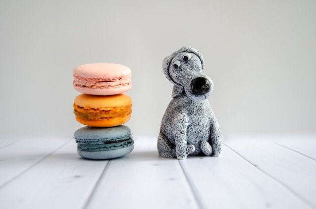 Cute funny toy dog looks at delicious sweet macaroons lying nearby. Concept: diet, avoiding sweets, proper nutrition, making confectionery. Humorous postcard, poster, space for text