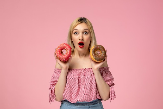 Cute girl adorable blonde chick with red lipstick in pink blouse shocked holding donuts