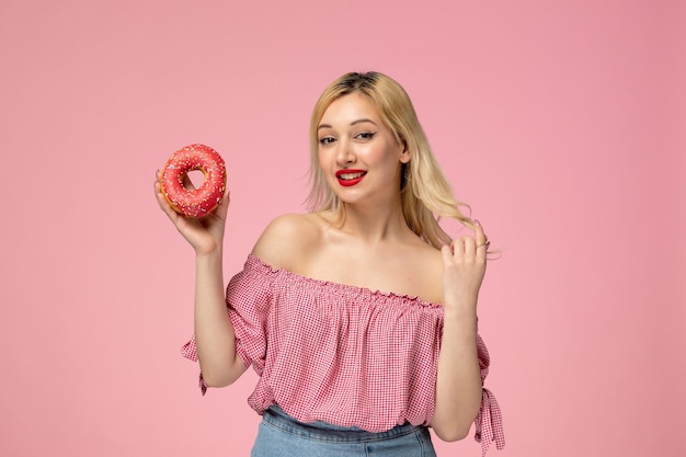 Cute girl adorable blonde chick with red lipstick in pink blouse touching hair and holding donut