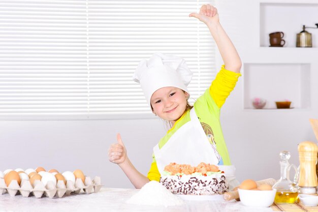 Ragazza carina che cuoce la torta in cucina che mostra i pollici in su