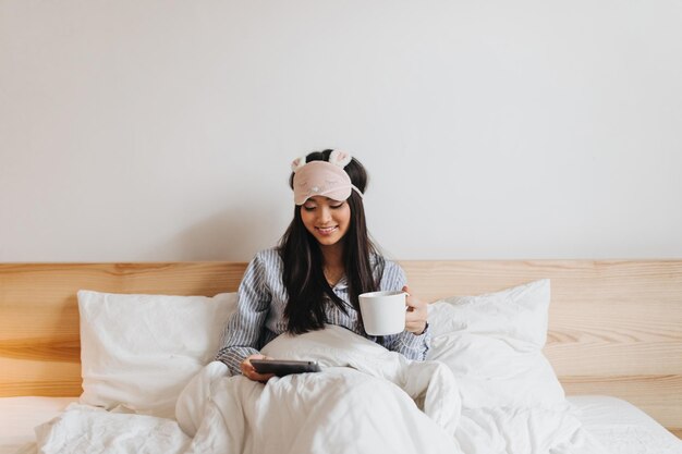 Ragazza carina in pigiama blu con sorriso legge sdraiato a letto con una tazza di tè