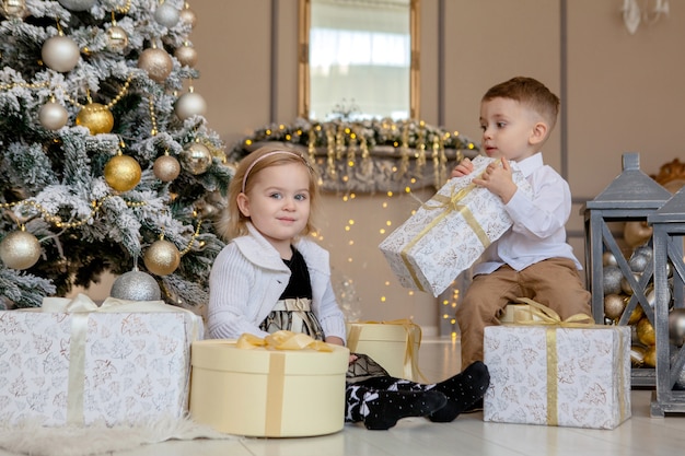 Ragazza carina e ragazzo che aprono i regali di natale. bambini sotto l'albero di natale con scatole regalo. soggiorno decorato con camino tradizionale. accogliente calda serata invernale a casa.