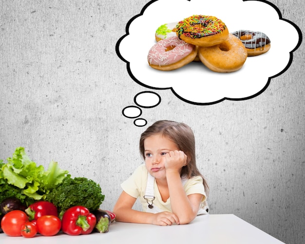 Cute girl child making a choice between healthy vegetables and unhealthy sugary donuts