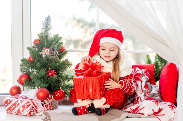 A cute girl child with a gift in her hands is sitting on the windowsill at the window of the house at the Christmas tree and waiting for the new year or Christmas in a red Santa Claus hat