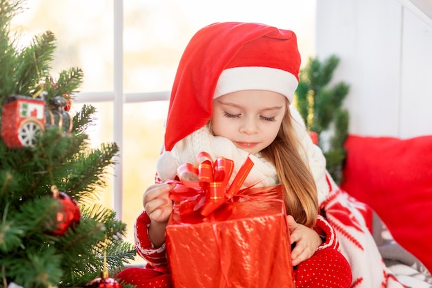 A cute girl child with a gift in her hands is sitting on the windowsill at the window of the house at the Christmas tree and waiting for the new year or Christmas in a red Santa Claus hat