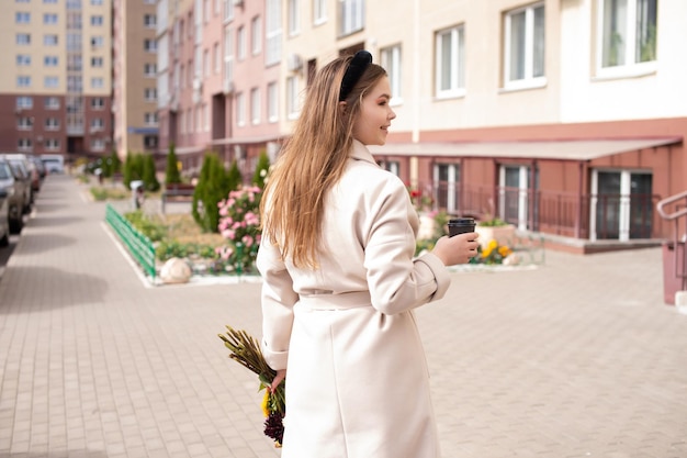 A cute girl in a coat holds a glass of coffee and a bouquet of flowers in the town