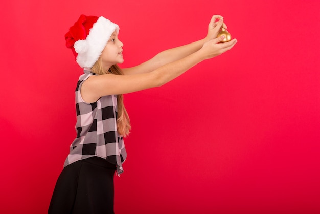 Cute girl on a colored background in a Christmas hat holds balls to decorate the Xmas tree - image