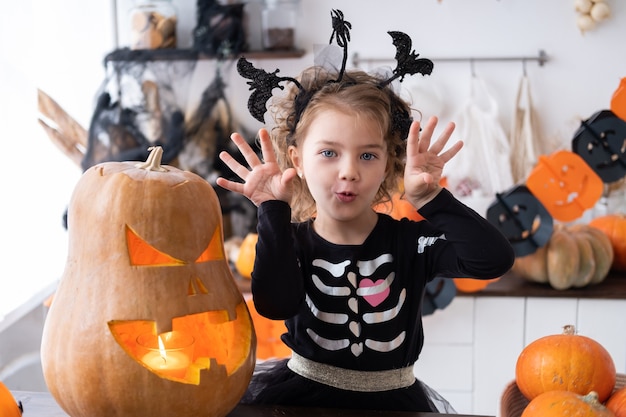 Cute girl in costume of witch with pumpkin at home in kitchen, having fun, celebrating Halloween.