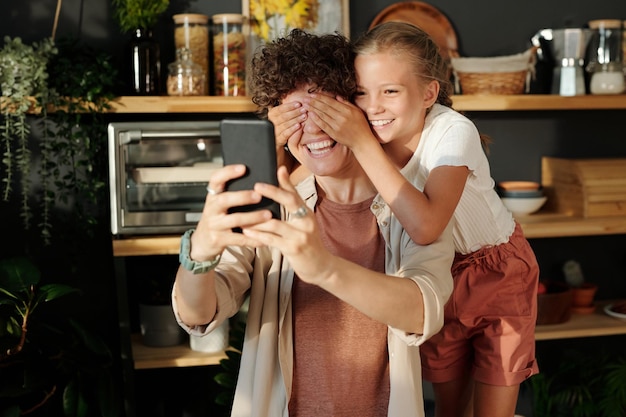 Ragazza carina che copre gli occhi di sua madre con le mani mentre si diverte durante il selfie