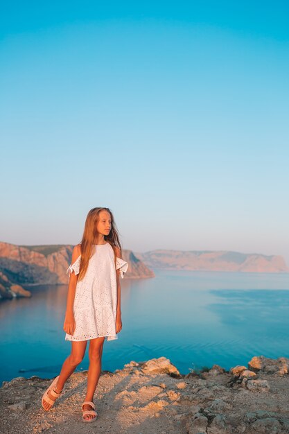 Cute girl in a dress on the mountain by the stunning rocks and sea at sunset. Beautiful nature