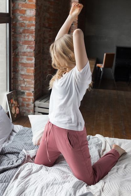 La ragazza sveglia si è vestita in pantaloni rosa del pigiama, maglietta bianca che si siede sul letto con il cuscino