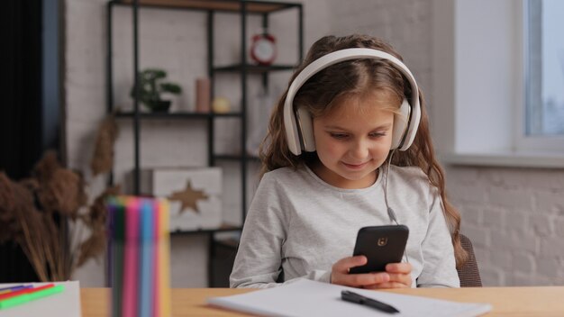 Cute girl in earphones studying at home using smartphone. Schoolgirl has online lesson.