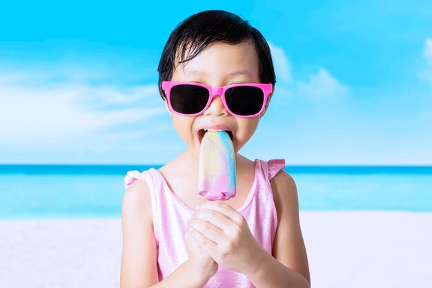 Cute girl eating popsicle while standing at beach against sky