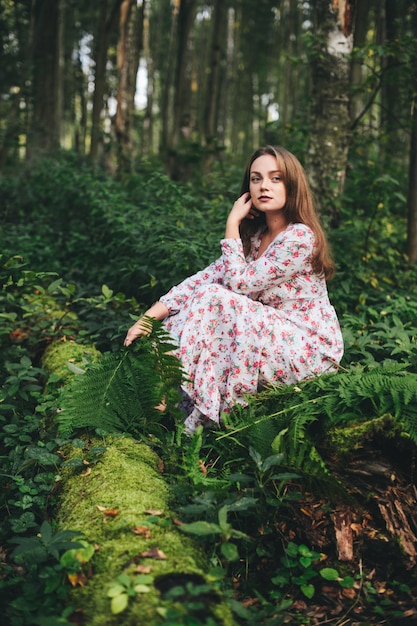 꽃 드레스에 귀여운 여자는 숲에서 고 사리 꽃다발과 함께 앉아있다.