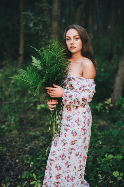 A cute girl in a floral dress is sitting with a fern bouquet in the forest. 