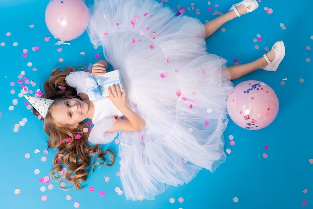 Cute girl in a fluffy white dress with a gift in her hands lies on a blue background in confetti