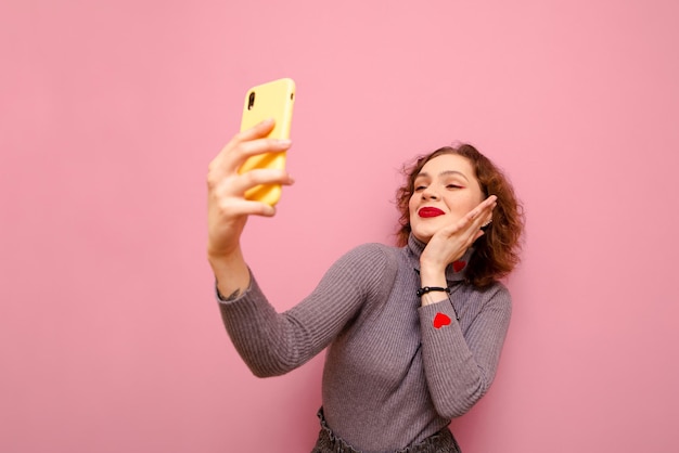 Cute girl in gray turtleneck on with curly red hair makes selfie on pink background