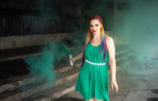 Cute girl in a green dress with African braids and colorful makeup posing with bright green smoke on the streets of a warm spring city