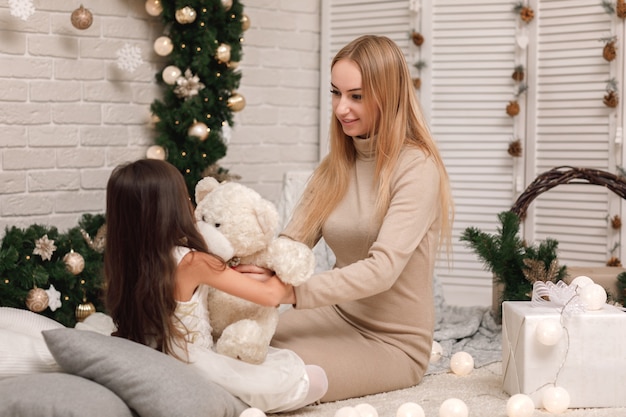 Cute girl and her mother playing with teddy bear near the Christmas tree at home