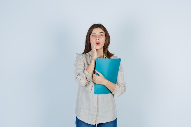 Cute Girl holding folder, keeping hand on cheek in shirt and looking surprised