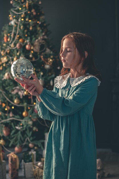 귀여운 소녀는 크리스마스 트리의 배경에 디스코 공을 들고 있습니다.