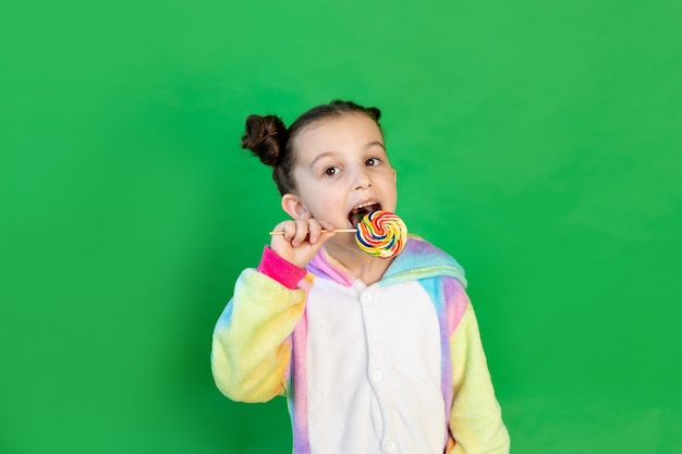 a cute girl is licking a large Lollipop in a bright suit on a green isolated background. Space for text.