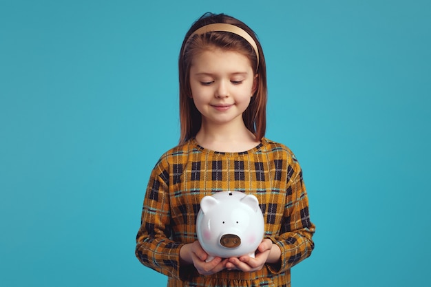 Cute girl kid holding money box looking down isolated over blue background