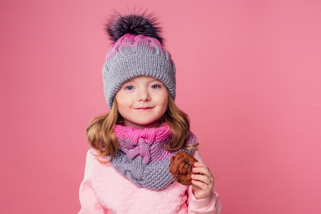 Cute girl in knitted hat eating Christmas biscuits.child model diet pink background in studio.