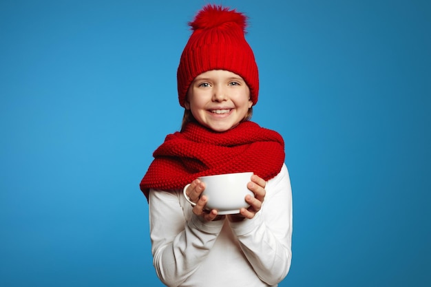 Cute girl in knitted red scarf and hat warming hands while holding cup of