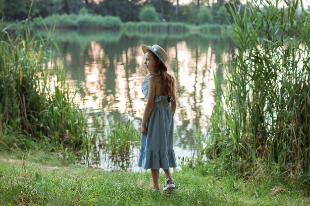 A cute girl in a long blue dress and straw hat stands on the shore of the lake. Back of a child in the public park with water, bushes, green and dried grass in summer. Young beautiful lady on walking.