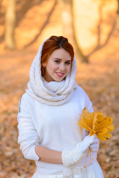 Ragazza carina in abito da sposa bianco lungo in posa in un percorso rurale tra alberi autunnali nella foresta nell'atmosfera dell'ora d'oro.